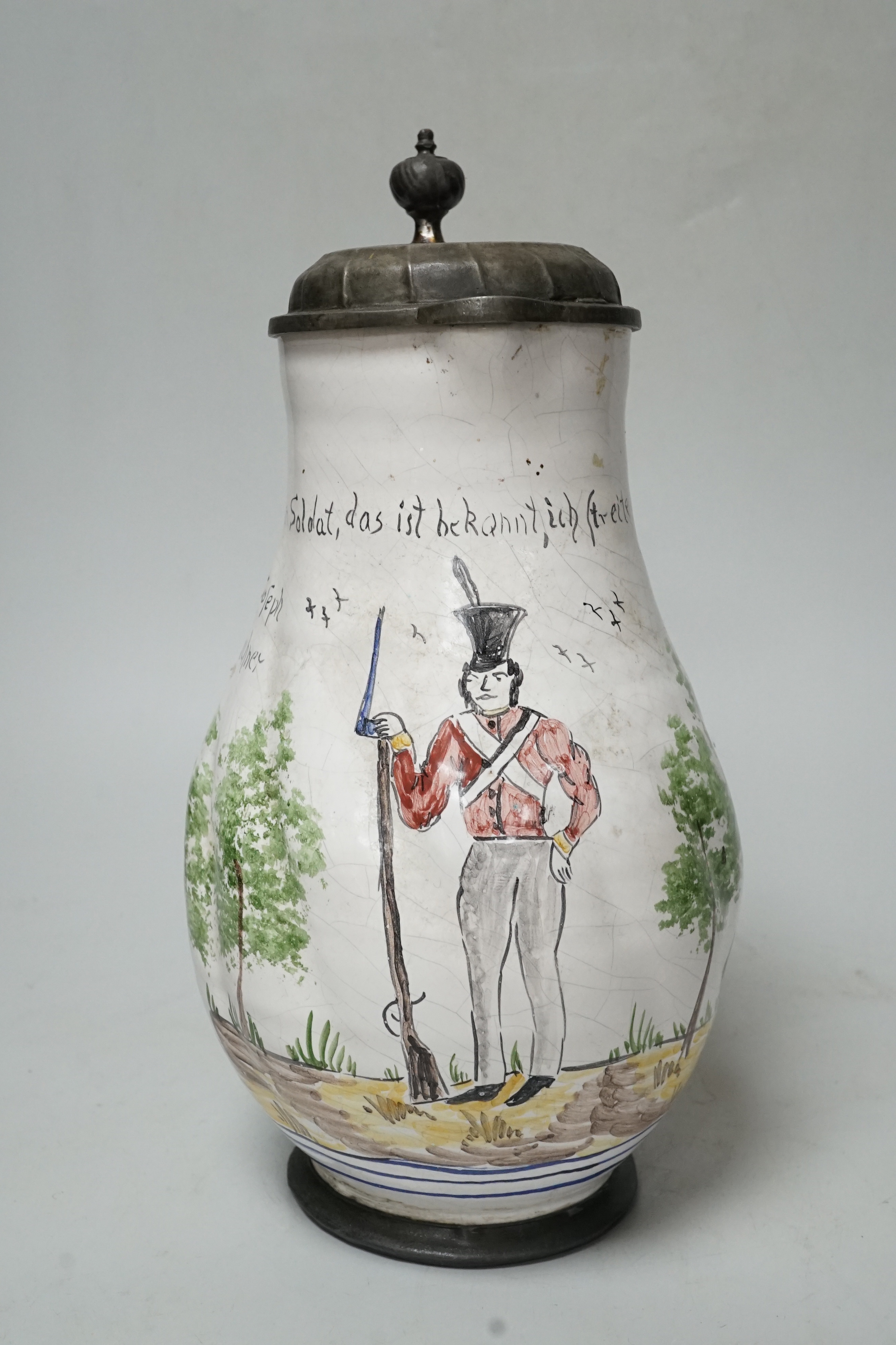 A large 18th century German faience pewter lidded jug, baluster shaped, with a naive painting of a soldier, inscribed ‘’Soldat, das ist bekqnnt, ich streite fur das vaterland’’ (Soldier, that is known for that fatherland